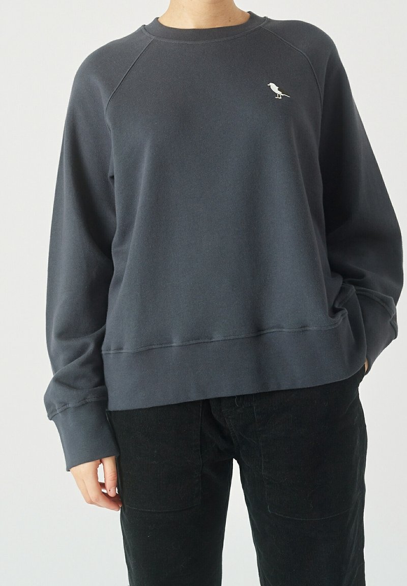 Felpa grigia con scollo a girocollo, maniche raglan e un piccolo logo di un uccellino bianco sul petto, abbinata a pantaloni in velluto a coste neri.