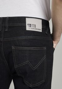 Jeans de mezclilla negra con un bolsillo trasero con costura en zigzag, parche de marca etiquetado como "TTD" y costuras en contraste a lo largo de las costuras.