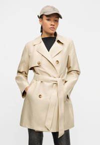 Νεαρό άτομο που φορά ένα μπεζ παλτό τρεντς με ζώνη, μαύρο τοπ, σκούρο παντελόνι και μπεζ καπέλο μπέιζμπολ, στεκόμενο με τα χέρια στις τσέπες.