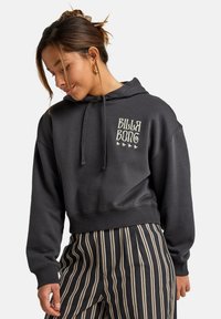 Sudadera negra recortada hecha de una tela suave, con un logo de colores claros y estrellas en el pecho, combinada con pantalones a rayas.