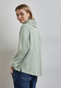 Ljusgrön, texturerad tröja med hög krage, långa ärmar och sidoflikar, som är matchad med mörkblåjeans.