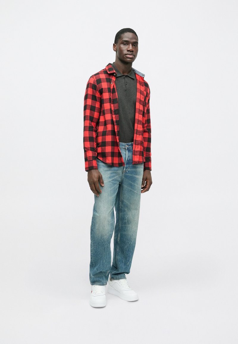 Red and black checkered flannel jacket over a dark polo, paired with light denim jeans and white sneakers; casual fit, textured fabric.