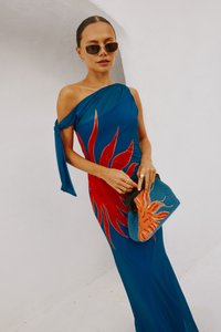 Una donna in un vestito blu a spalla unica con un grande disegno di sole rosso, mentre tiene una borsa a pochette blu e arancione a tema sole contro un muro bianco.