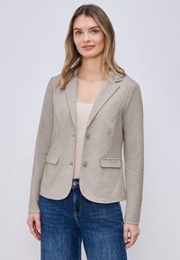 Femme portant un blazer beige clair cintr é par-dessus un haut crème et un jean bleu, debout devant un fond gris clair uni.