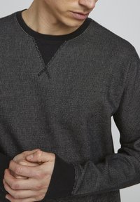 Sweat-shirt noir à motifs présentant un design texturé, avec un col en V et des poignets noirs, tenu par un mannequin avec les mains jointes.
