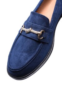 Mocassini in suede blu navy con suola piatta in gomma, caratterizzati da un accento decorativo in catena argentata sulla parte anteriore, finitura texture e linee pulite.