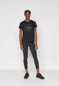 Čierne tričko DKNY s trblietavým logom, v kombinácii s tmavohnedými legínami. Modelka má na sebe čierne športové topánky a je pózovaná na jednoduchom bielom pozadí.