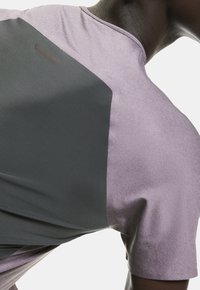 Lyhythihainen urheilupaita vaaleanvioletissa ja tummanharmaassa värissä. Ominaisuuksiin kuuluu teksturoitu kangas, rento istuvuus ja minimalistinen logoelementti takana.