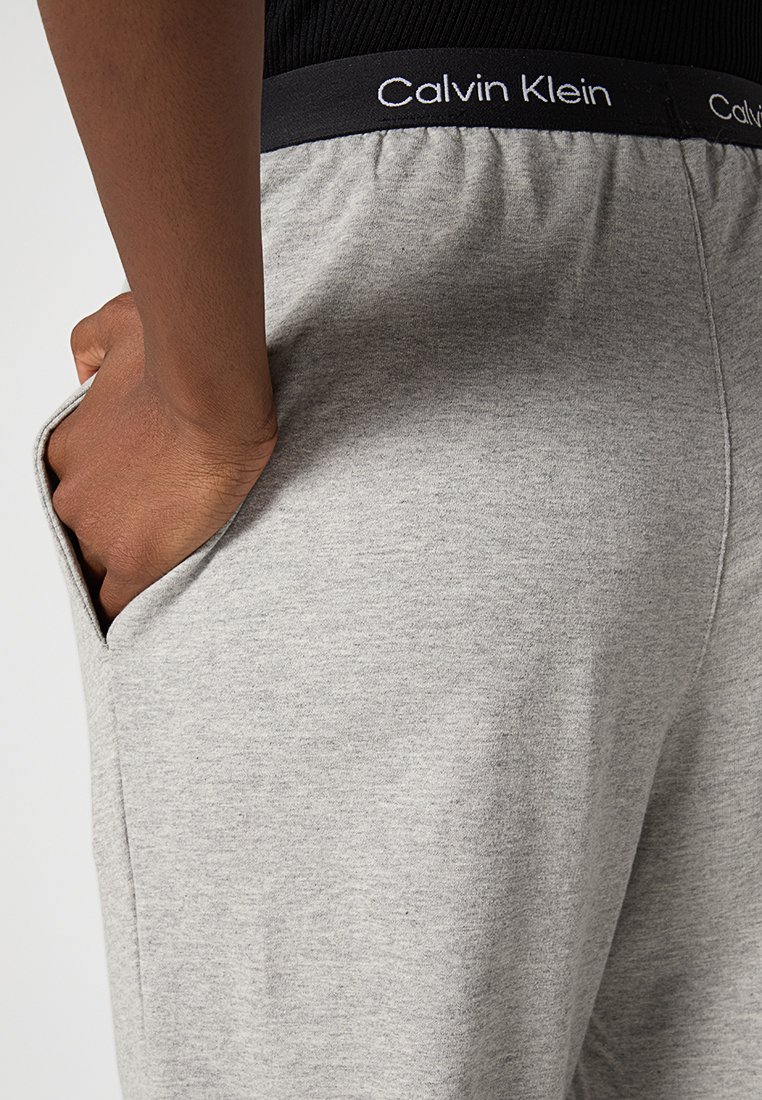 Mano nella tasca di pantaloni della tuta grigio chiaro con cintura nera etichettata "Calvin Klein," indossati con un top nero.