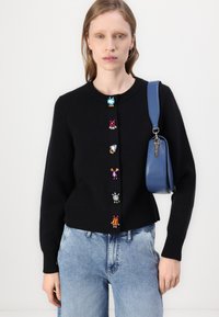 Čierny cardigan s farebnými kamienkovými gombíkmi na prednej strane, s okrúhlym výstrihom a rebrovanými manžetami, skombinovaný so svetlomodrými džínsami a modrou taškou.