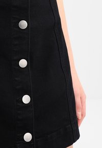 Schwarzer Denim-Rock mit silbernen Druckknöpfen an der Vorderseite, taillierte Passform und glatte Textur. Eine teilweise sichtbare Hand an der Seite.