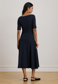 Donna che indossa un abito midi blu navy con maniche a tre quarti, scarpe slingback nere e braccialetti dorati, in piedi su un pavimento di legno.