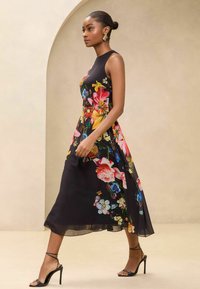 Schwarzes Blumenkleid mit tailliertem Oberteil, fließendem Saum und lebhaften, mehrfarbigen Blumenmustern; kombiniert mit schwarzen, geschnürten Absatzschuhen.