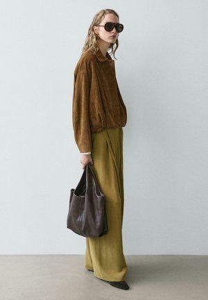 Vrouw draagt een bruine suède jas, olijfgroene broek met wijde pijpen, zwarte zonnebril, houdt een grote donkerbruine leren tas vast en staat tegen een effen muur.