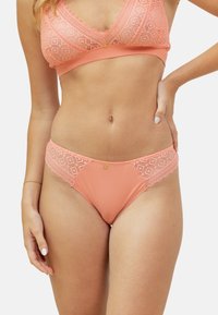 Sous-vêtements avec des bordures en dentelle en tissu doux pêche, présentant un motif géométrique à l'avant et une texture lisse avec un détail d'accent doré.