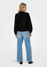 Donna bionda di spalle, che indossa una blouse nera a maniche lunghe, jeans larghi blu chiaro e stivali neri con tacco su uno sfondo neutro.