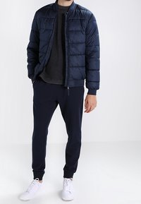 Giacca puffer blu navy con zip frontale e polsini a costine, indossata sopra una t-shirt scura e pantaloni jogger blu navy abbinati, completata da sneakers bianche.