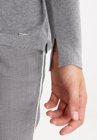 Grauer langärmeliger Pullover mit gerippter Textur und seitlichen Schlitzen, kombiniert mit schwarz-weiß karierten Hosen. Nahaufnahme einer Hand.