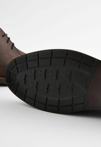 Suela de zapato de cuero marrón con una parte inferior de goma negra texturizada. Presenta patrones de tracción triangulares y un diseño de borde suave.