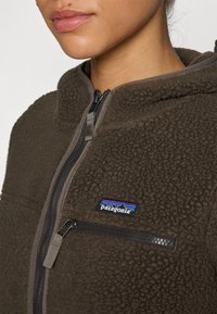Brun fleecejakke med høy krage, glidelås foran og brystlomme. Har et Patagonia-logo med blå og røde detaljer.