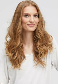 Langes, welliges, hellbraunes Haar umrahmt ein Gesicht. Sie trägt ein lockeres, cremefarbenes Oberteil. Der Hintergrund ist neutral und sanft.