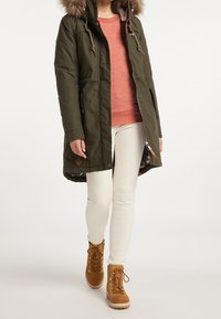 Olivgrön parka med pälsfodrad huva, knappdetaljer och dragsnören. Bärs över en texturerad persikofärgad tröja, vita byxor och bruna stövlar.