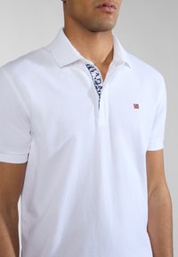 Uomo che indossa una polo bianca a maniche corte con piccola bandiera norvegese sul petto e colletto a pois blu navy.