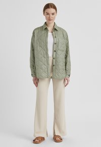 Grüne gesteppte Jacke mit Kragen, Knopfverschluss und vorderer Tasche, kombiniert mit cremefarbenen weiten Hosen und hellbraunen Sandalen.