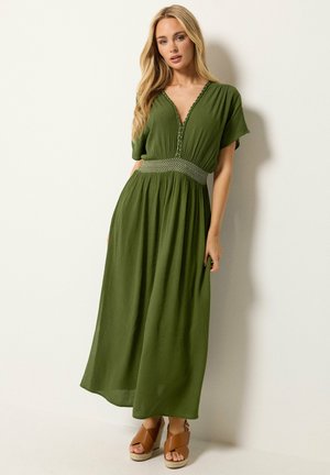 Vrouw met lang blond haar, die een groene maxi-jurk met V-hals en korte mouwen en bruine sleehak sandalen draagt, staande tegen een witte muur.