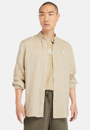 Camicia button-up in lino beige con colletto, maniche arrotolate e bottoni bianchi. Indossata sopra una t-shirt grafica color nocciola, abbinata a pantaloni verde oliva.