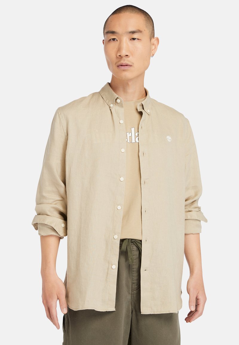 Beige linnen overhemd met knopen, een kraag, opgerolde mouwen en witte knopen. Gedragen over een tan grafisch T-shirt, gecombineerd met olijfgroene broek.