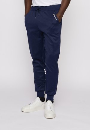 Mørkeblå sweatpants med elastisk linning og snøre, forsynet med sidelommer og hvid logo accent. Materialet fremstår blødt og glat.