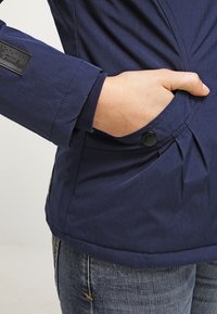Giacca blu navy con una finitura testurizzata, caratterizzata da una tasca cucita e dettagli con bottoni. Include un polsino con un accento in maglia.