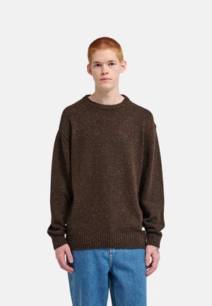 Jeune personne aux cheveux courts et roux portant un pull ample marron moucheté et un jean bleu, debout face à un fond clair uni.