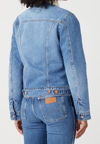 Chaqueta de denim azul claro con un diseño entallado, detalles de botones y una textura ligeramente desgastada. En la parte posterior lleva una pequeña etiqueta de la marca.