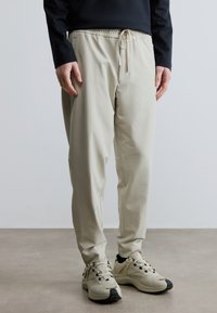 Világos bézs színű, rugalmas derékpánttal ellátott joggingnadrág, amelyen húzózsinór, oldalzsebek és szűkített szárú kialakítás található. Semleges színű tornacipővel viselve.