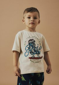 Camiseta color beige claro con un gráfico de dinosaurio, letras azules y un diseño de patineta. Se combina con pantalones de pijama oscuros con patrones de dinosaurios.
