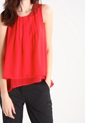 Femme portant un chemisier rouge sans manches à volants avec un col décoratif et un pantalon noir, debout avec une main dans la poche.