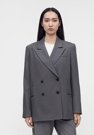 Femme aux longs cheveux noirs porte un blazer gris oversize à double boutonnage sur un haut blanc et un pantalon à carreaux, debout devant un fond clair uni.