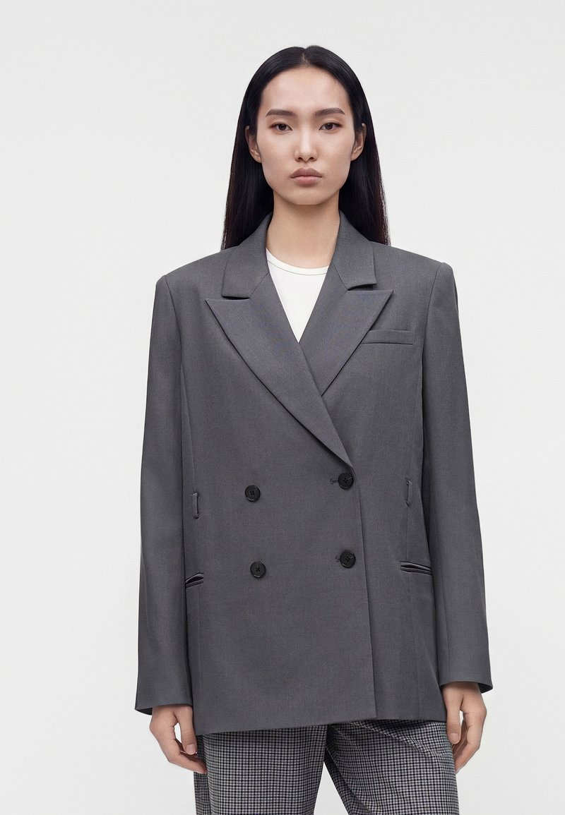 Femme aux longs cheveux noirs porte un blazer gris oversize à double boutonnage sur un haut blanc et un pantalon à carreaux, debout devant un fond clair uni.