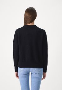 Schwarzer Strickpullover mit runder Halslinie, langen Ärmeln und lockerer Passform. Weicher Stoff, getragen über hellblauen Jeans.