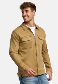 Beige bomuldsjakke med krave, der har to brystlommer, en lynlås foran og lange ærmer. Båret over en hvid skjorte med denimjeans.
