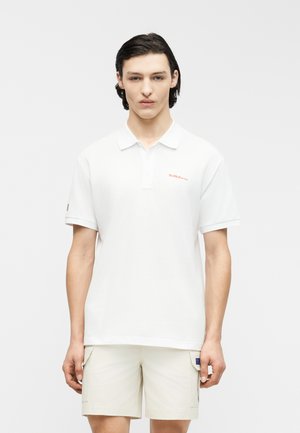 Jeune homme aux cheveux foncés portant un polo blanc avec le logo Red Bull Racing et un short cargo beige clair, debout devant un fond uni.
