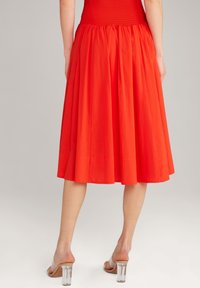 Jupe rouge jusqu'aux genoux avec une taille smockée. Fabriquée en tissu léger, avec un design fluide et plissé. Sandales à talons blocs transparentes visibles.