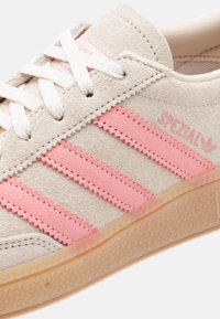 Ανοιχτό μπεζ σουετ αθλητικό παπούτσι με ροζ δερμάτινες ρίγες, λευκά κορδόνια και λαστιχένια σόλα. Διαθέτει υφασμένο μοτίβο και λογότυπο στην πλευρά.