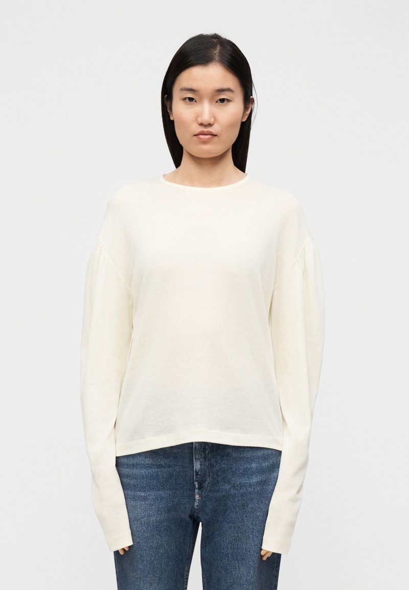 Kremasta majica z dolgimi rokavi z okroglim izrezom, sproščenim krojem in mehko teksturo. Kombinirana s temno modrimi oprijetimi kavbojkami. Preprost dizajn, brez vzorcev.