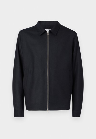 Melna garām piedurknēm jaka ar apkakli un centrālu sudraba rāvējslēdzēju, kas aprīkota ar divām sānu kabatām, attēlota uz balta fona.