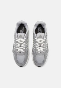 Chaussures de sport grises avec une tige en maille, trois rayures gris clair et des lacets blancs. La semelle est principalement blanche avec des accents texturés.