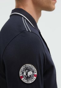 Marineblaues Poloshirt mit weißen Akzentnähten, versehen mit einem kreisförmigen, gestickten Abzeichen auf der linken Schulter, das eine Weltkugel und Textdetails zeigt.