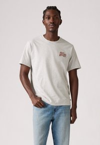 T-shirt de algodão cinza com decote redondo e mangas curtas, apresentando um logotipo castanho na área esquerda do peito. Combinado com jeans azul claro.
